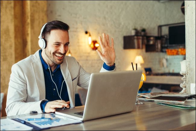 A man wearing headphones and waving at his laptop.